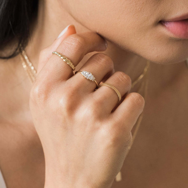 Second Info Card, A woman's hand adorned with elegant gold rings, including a twisted band, a delicate diamond ring, and a thin textured band.