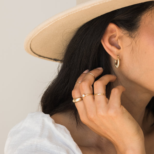 Third Info Card, A woman wearing gold hoop earrings and layered rings, including a pearl accent ring, while styling her hair with a beige hat.