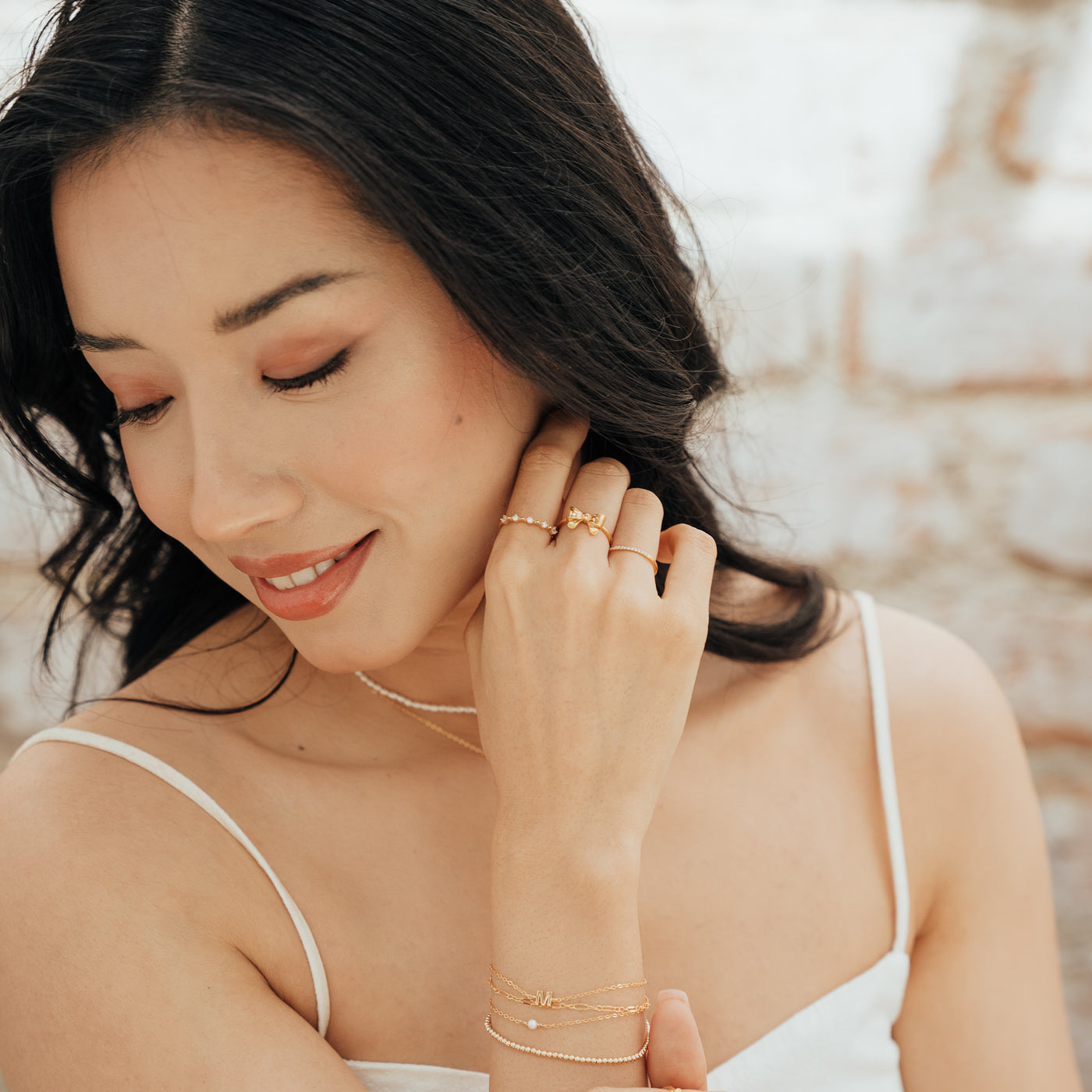 Woman looking down to the side with her hand near her face, wearing a CZ Floating Ring, Ribbon Bow Ring, Dainty Ring, Stretch Bead Bracelet, Freshwater Pearl Bracelet, and Initial Bracelet.