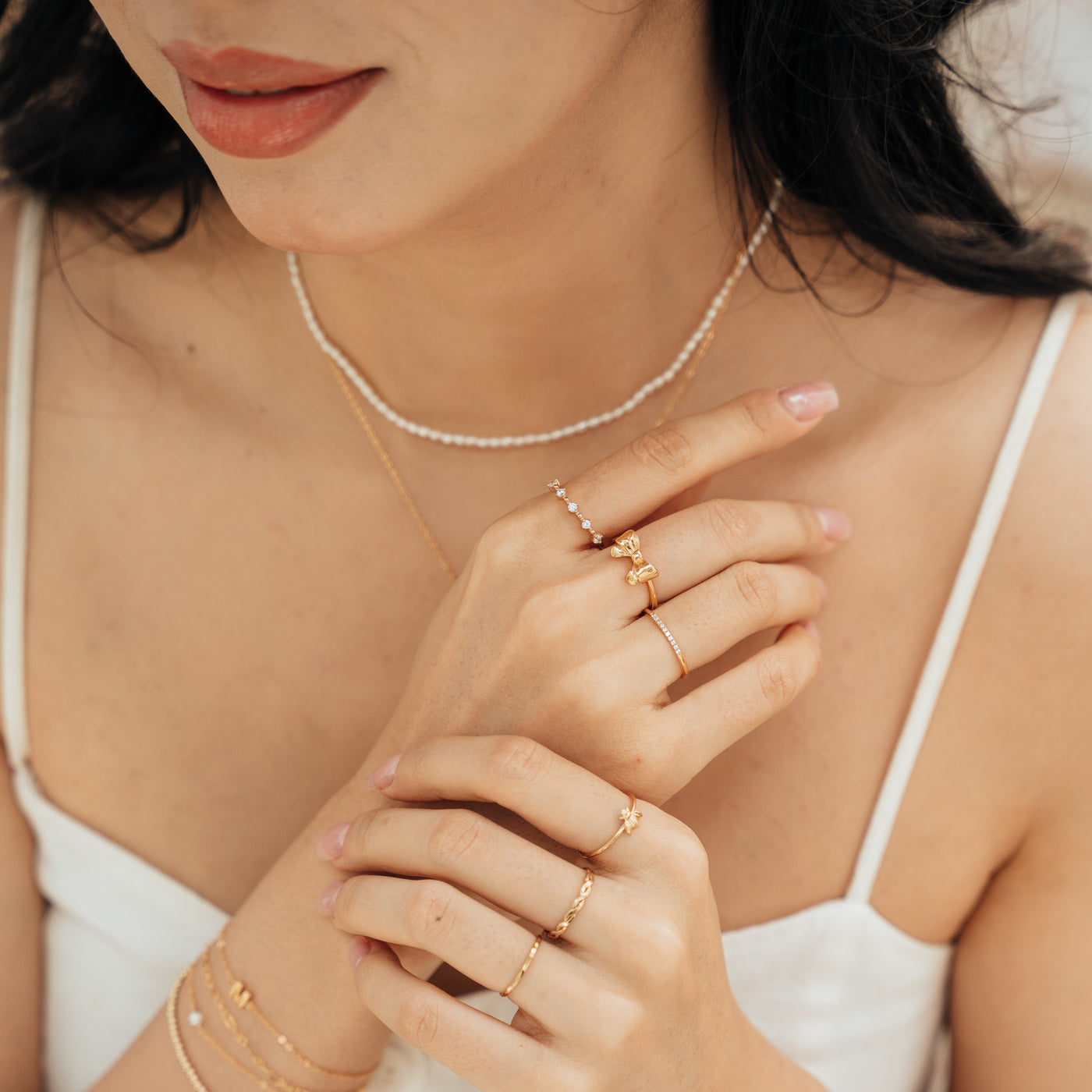 Close-up of a woman’s chest, gently holding one wrist with her other hand, wearing jewelry including a Floating CZ Ring, Ribbon Bow Ring, Pavé Ring, Bee Ring, Twisted and Dapped Ring, and a Pearl Strand Necklace.