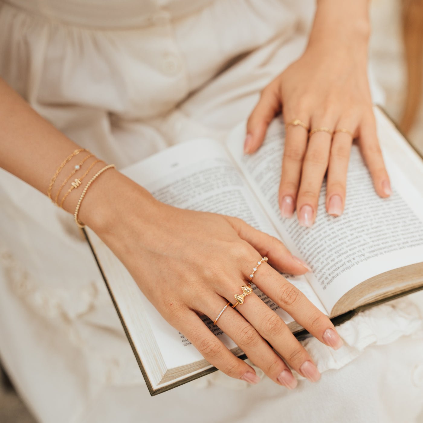 Close-up of a woman’s lap with a book resting on it and her hands placed on the book, wearing jewelry including a Bow Ring, Pavé Ring, CZ Floating Ring, and several Bracelets.