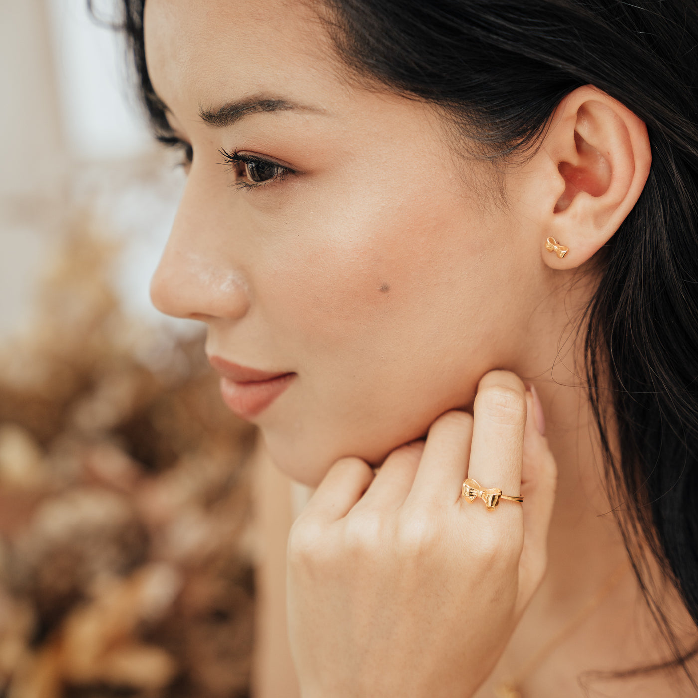 Close-up of a woman resting her face on her hand, wearing a gold vermeil Ribbon Bow Ring on her index finger and matching Ribbon Bow Stud Earrings