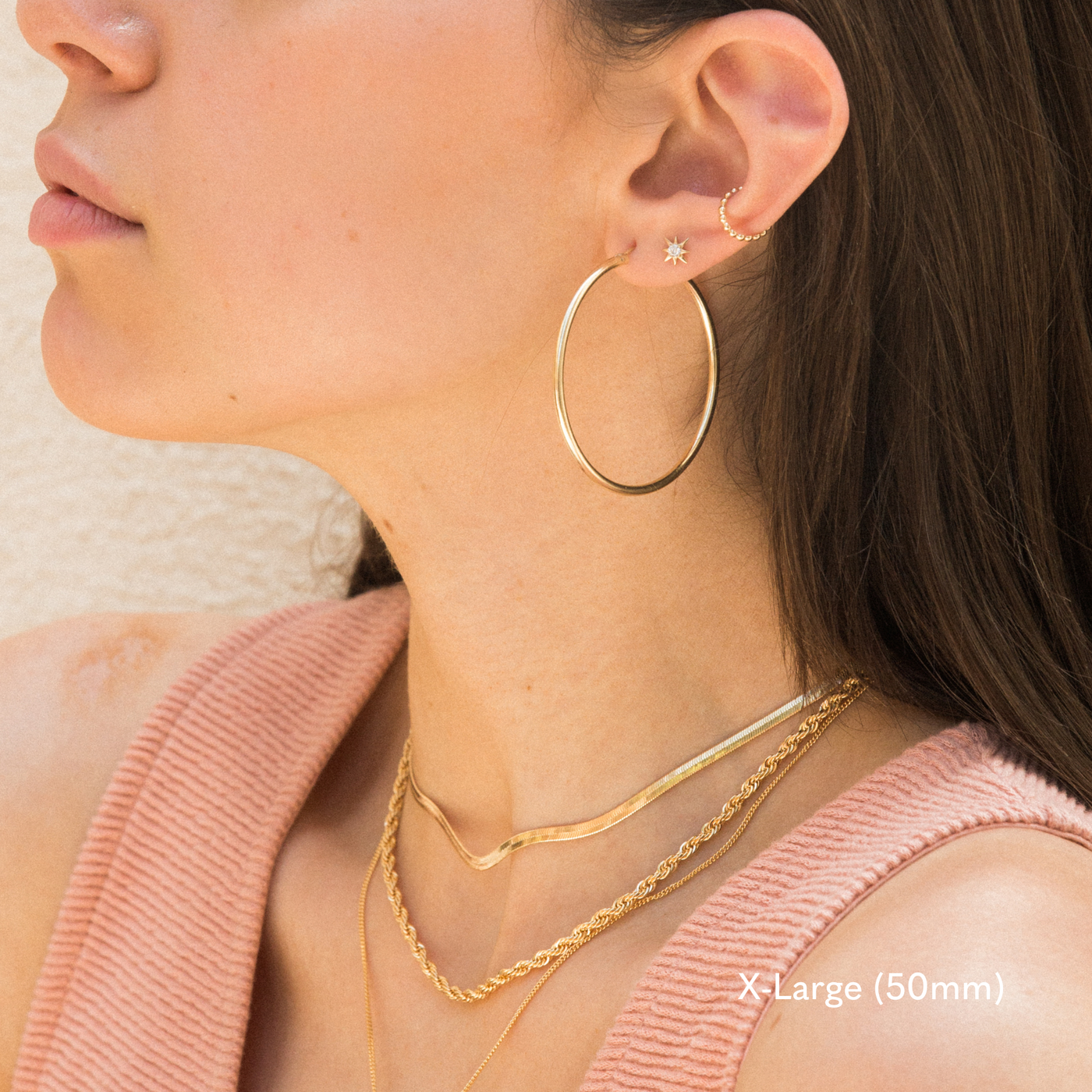 Side profile of a woman wearing an extra large 50mm gold hoop earring, small star stud, and gold ear cuff. She’s dressed in a pink textured tank top and layered gold necklaces. The image is taken in natural light against a light-colored wall.

X-Large (50mm), Everyday Hoop Earrings by Simple & Dainty Jewelry