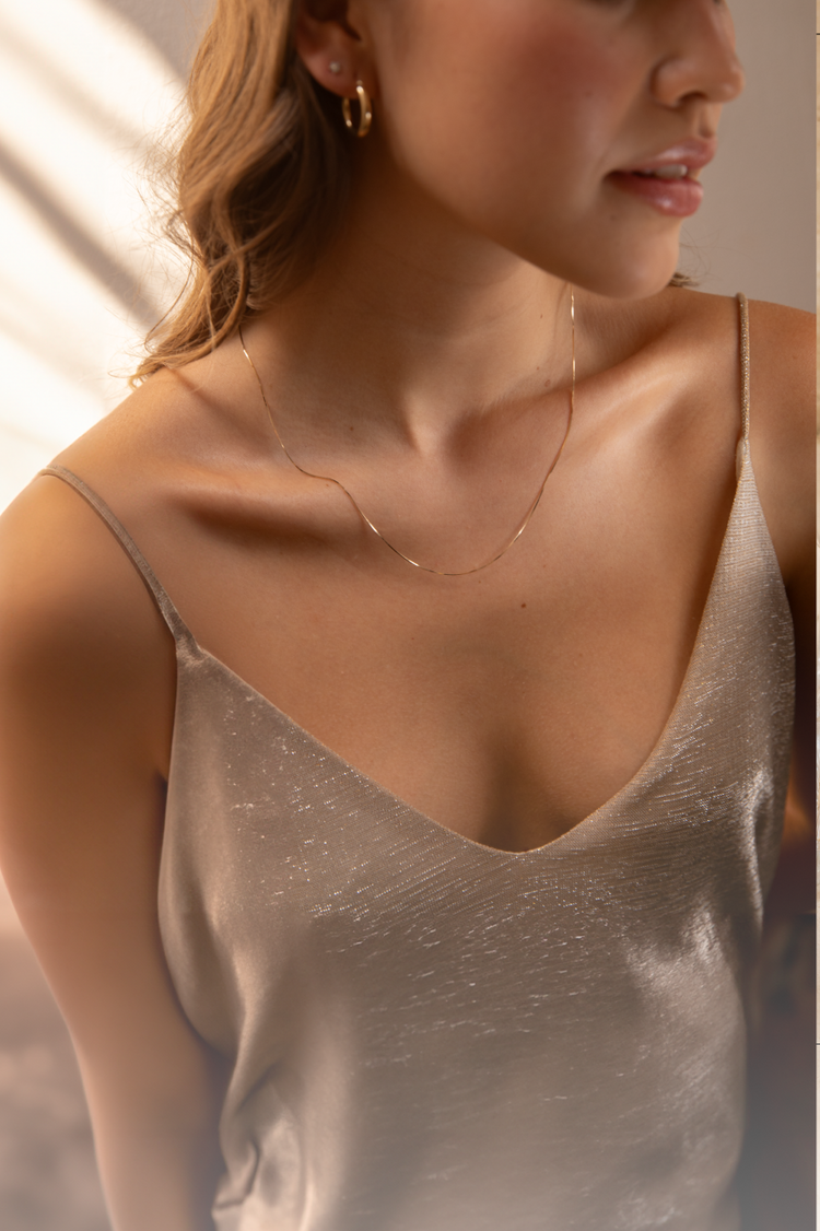 Close-up of a woman wearing a delicate gold necklace and hoop earrings, with a soft beige background and sunlight shadows.