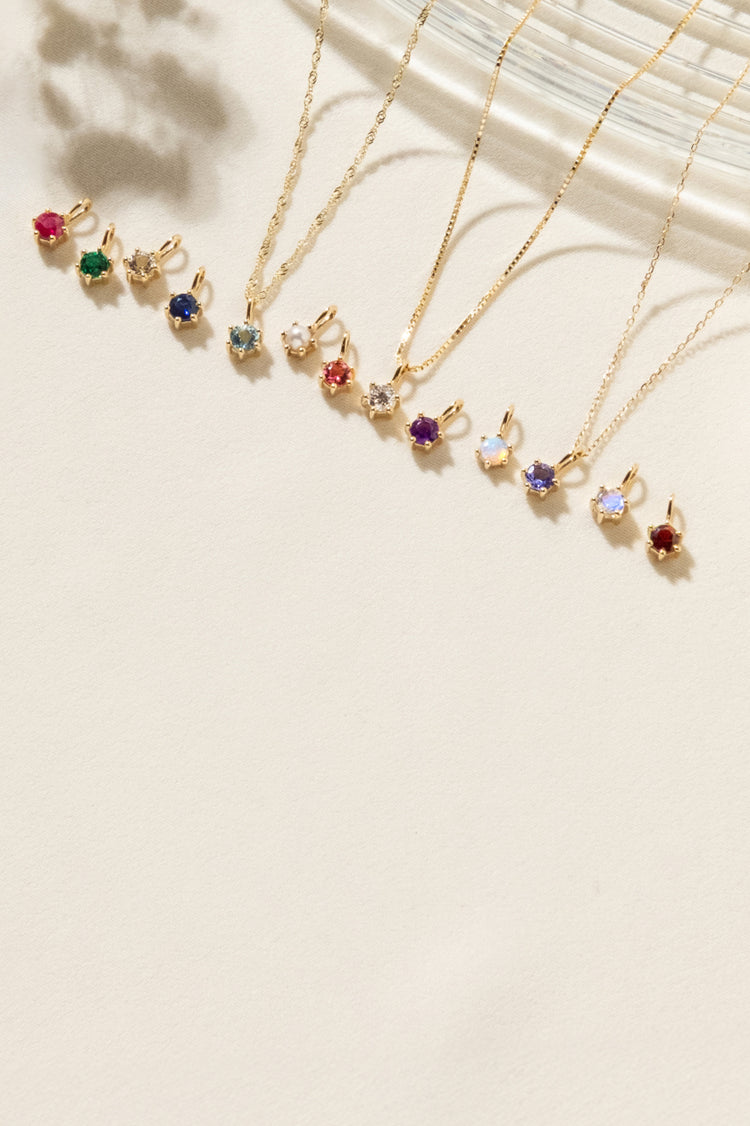 14k solid gold birthstone charms lying on light surface including: Ruby, emerald, diamond, sapphire, aquamarine, pearl, tourmaline, topaz, amethyst, opal, moonstone, and garnet.