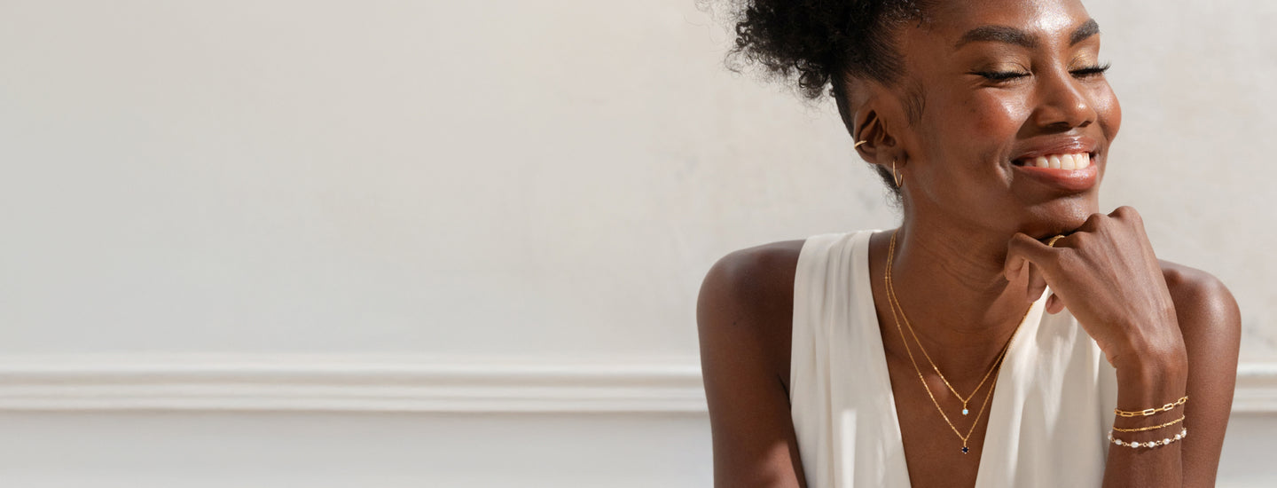 Woman smiling with her hand on her chin, wearing layered jewelry including two birthstone necklaces, three bracelets, hoop earrings, and an ear cuff.