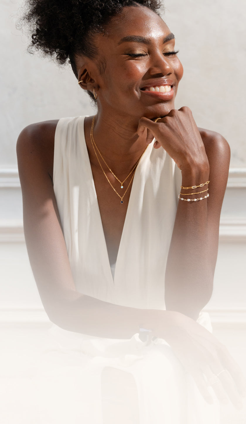 Woman smiling with her hand on her chin, wearing layered jewelry including two birthstone necklaces, three bracelets, hoop earrings, and an ear cuff.