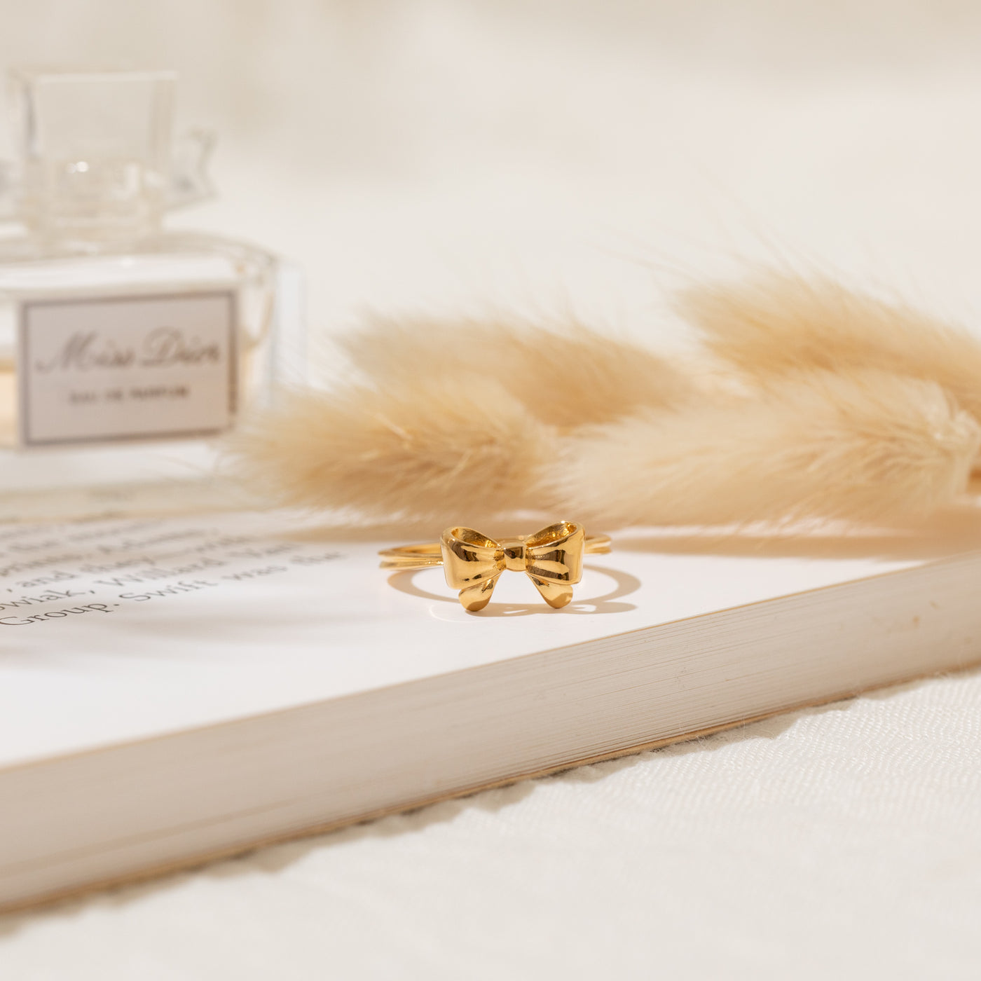 Gold vermeil Ribbon Bow Ring resting on an open book, with pampas grass and a perfume bottle blurred in the background.