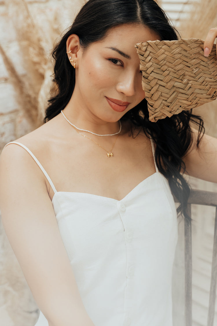Woman partially covering her face with a natural fiber fan, wearing a pearl necklace layered with a bow necklace, plus matching stud earrings and rings.
