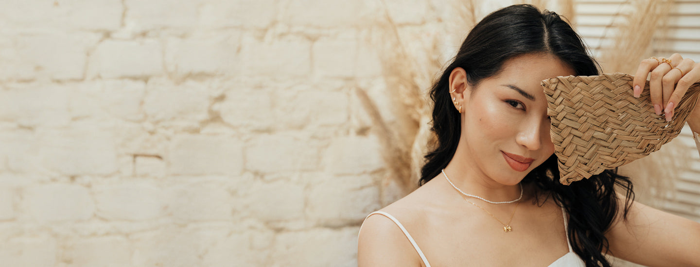 Woman partially covering her face with a natural fiber fan, wearing a pearl necklace layered with a bow necklace, plus matching stud earrings and rings.
