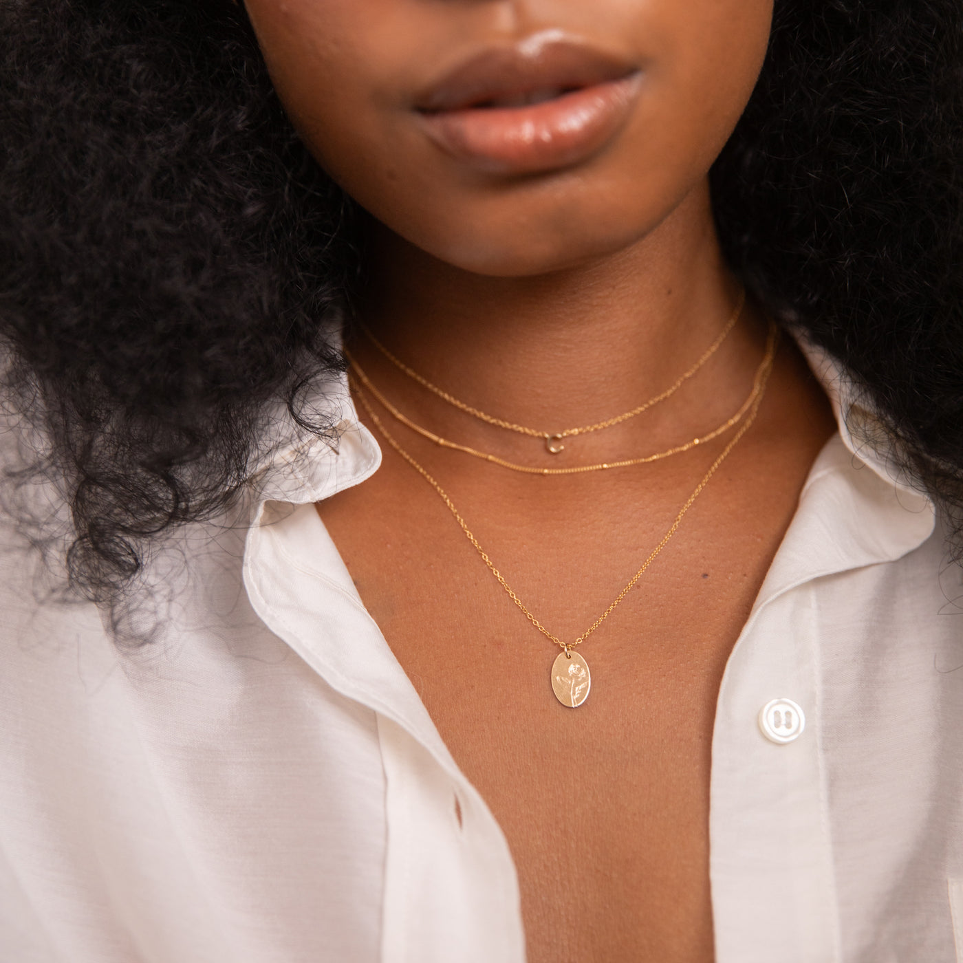 Woman wearing three layered gold necklaces: Tiny Initial Necklace, Dainty Satellite Chain Necklace, and Birth Flower Necklace. She’s in a white button-up shirt with the top buttons undone.
