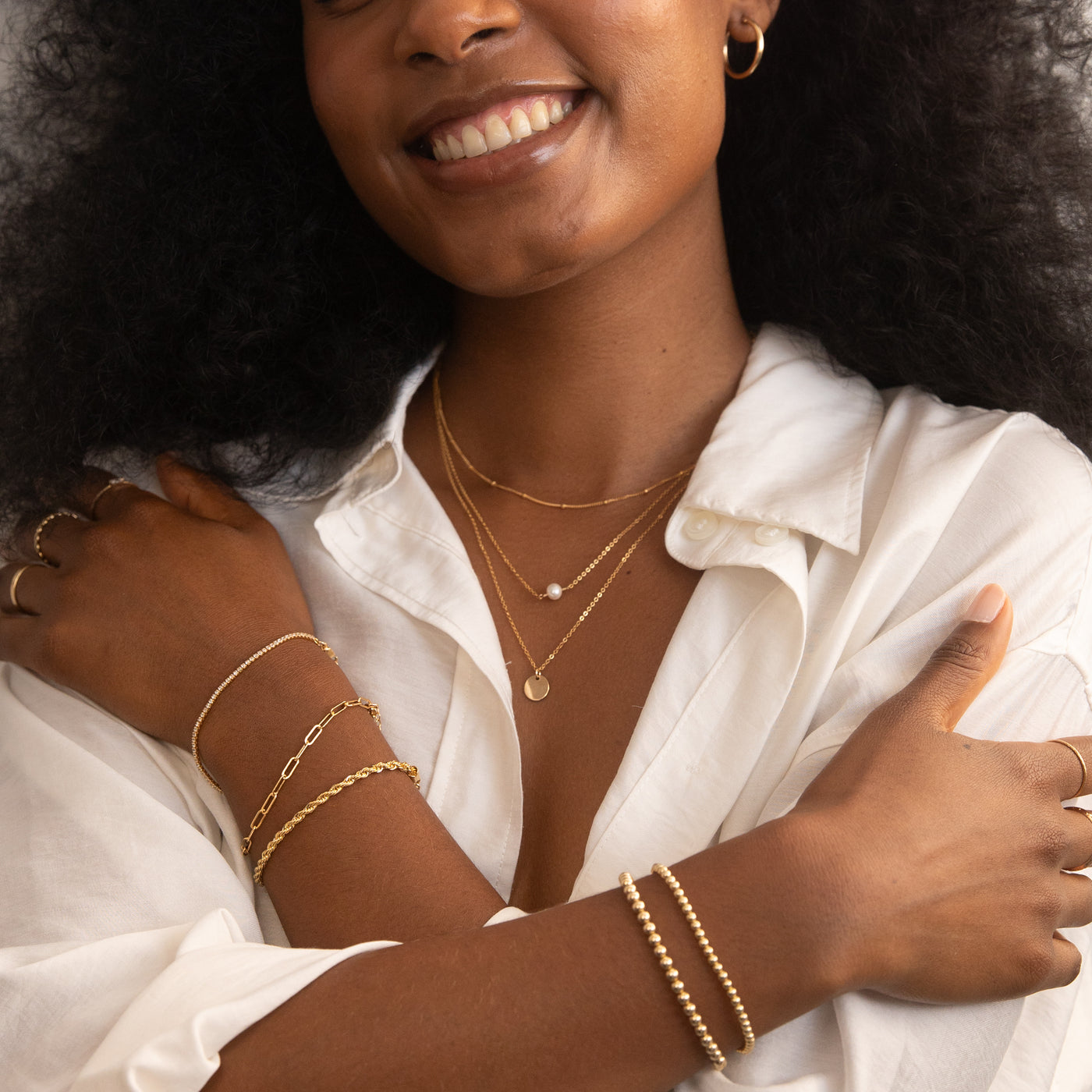 Woman wearing the Satellite & Coin Necklace Set and Freshwater Pearl Necklace, along with gold bracelets: Dainty CZ Tennis Bracelet, Chunky Paperclip Bracelet, and French Rope Chain Bracelet on the left wrist, and a Stretch Bead Bracelets on the right. She is dressed in a white button-up shirt, smiling with arms crossed to showcase the jewelry.