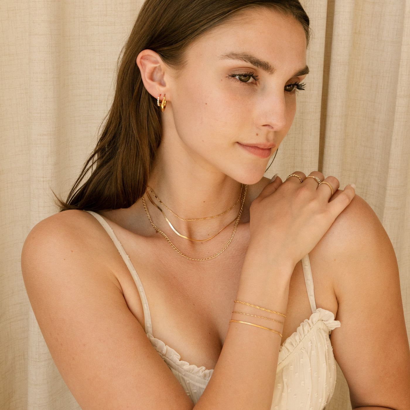 Woman looking to the side and grabing her shoulder with her arm. She's wearing chain necklaces, bracelets, rings, and hoop earrings.