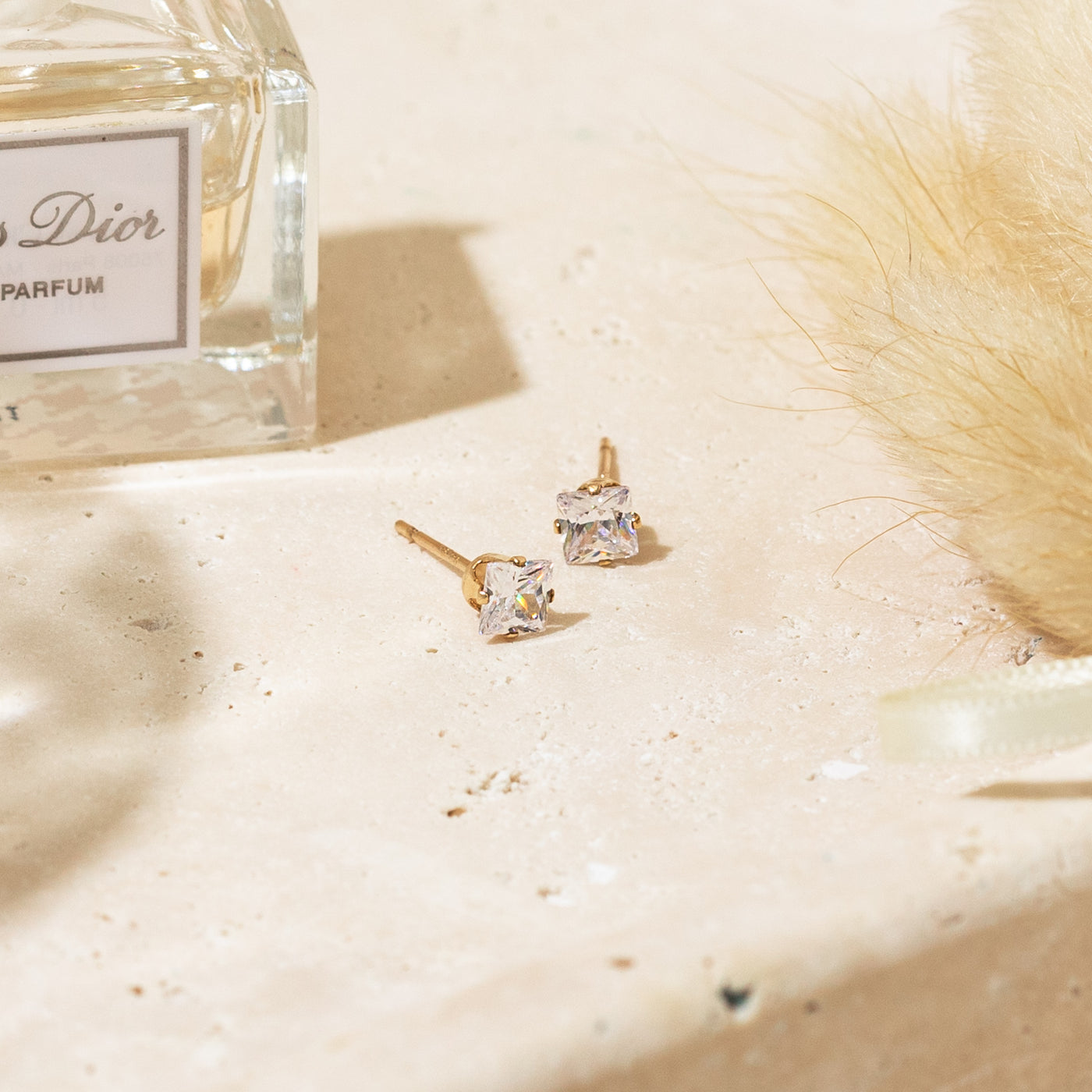 3mm 4mm Dainty Square CZ Stud Earrings. Gold stud earrings on a textured beige surface with a Dior perfume bottle in the background.