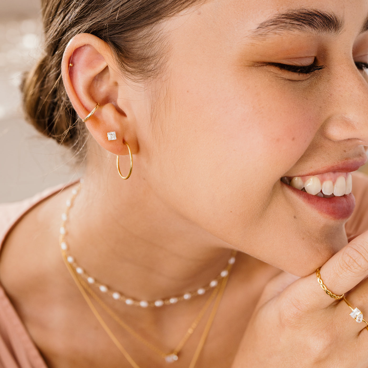 3mm 4mm Dainty Square CZ Stud Earrings. Close-up of a woman wearing gold hoop earrings, necklace, and ring with a blurred background.