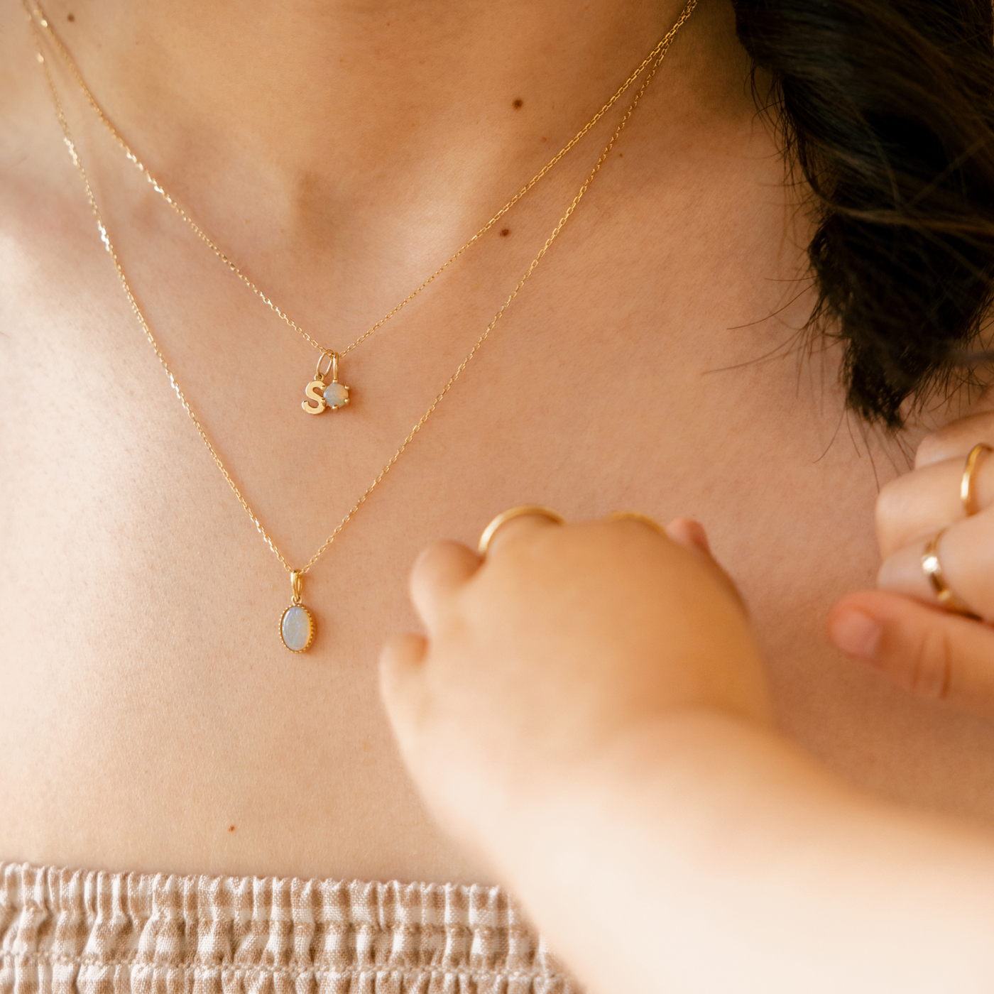 Close-up of a person wearing gold necklaces and rings with a soft background. A B C D E F G H I J K L M N O P Q R S T U V W X Y Z Tiny Hanging Initial Necklace