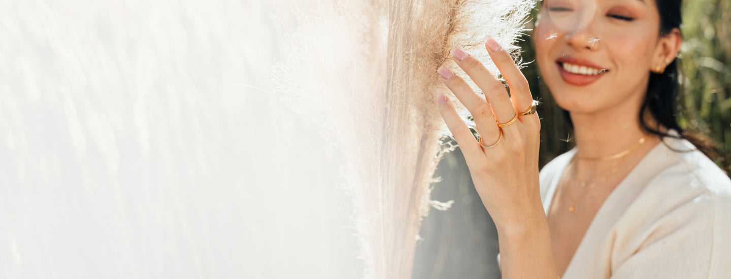 Woman outdoors touching pampas grass while wearing three gold rings.
