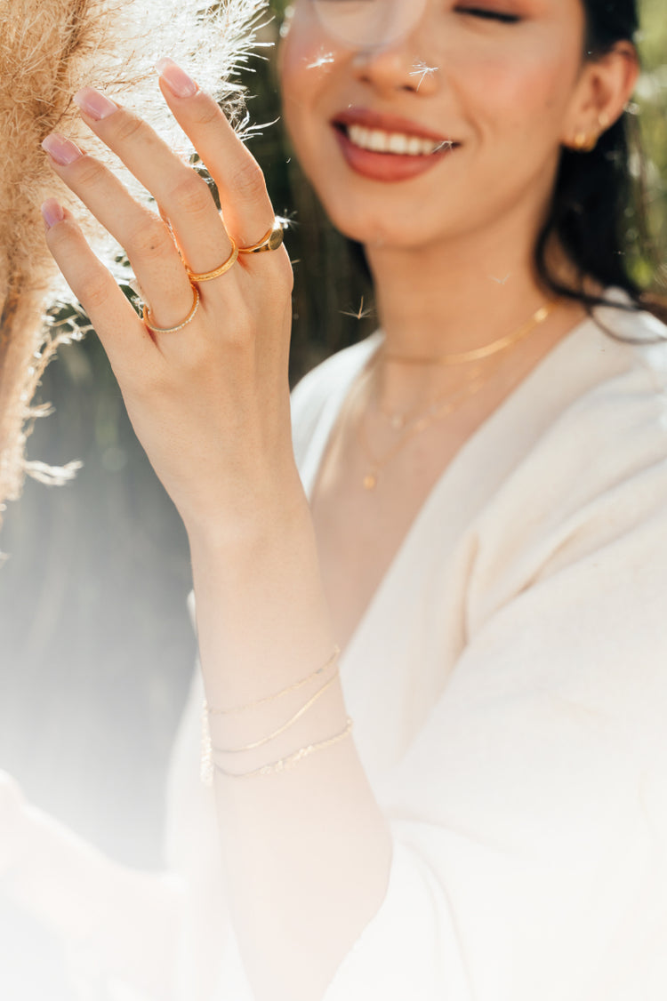 Woman outdoors touching pampas grass while wearing three gold rings.