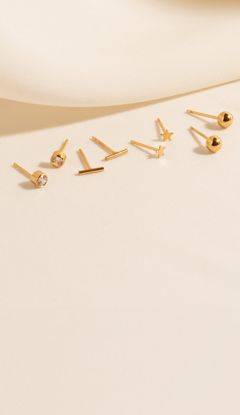 An image of six stud earrings against a cloth background: Dainty CZ Bezel Stud, Tiny Bar Studs, Tiny Star Studs, 4mm Ball Studs, 2mm Ball Studs, and Bead Bar Studs