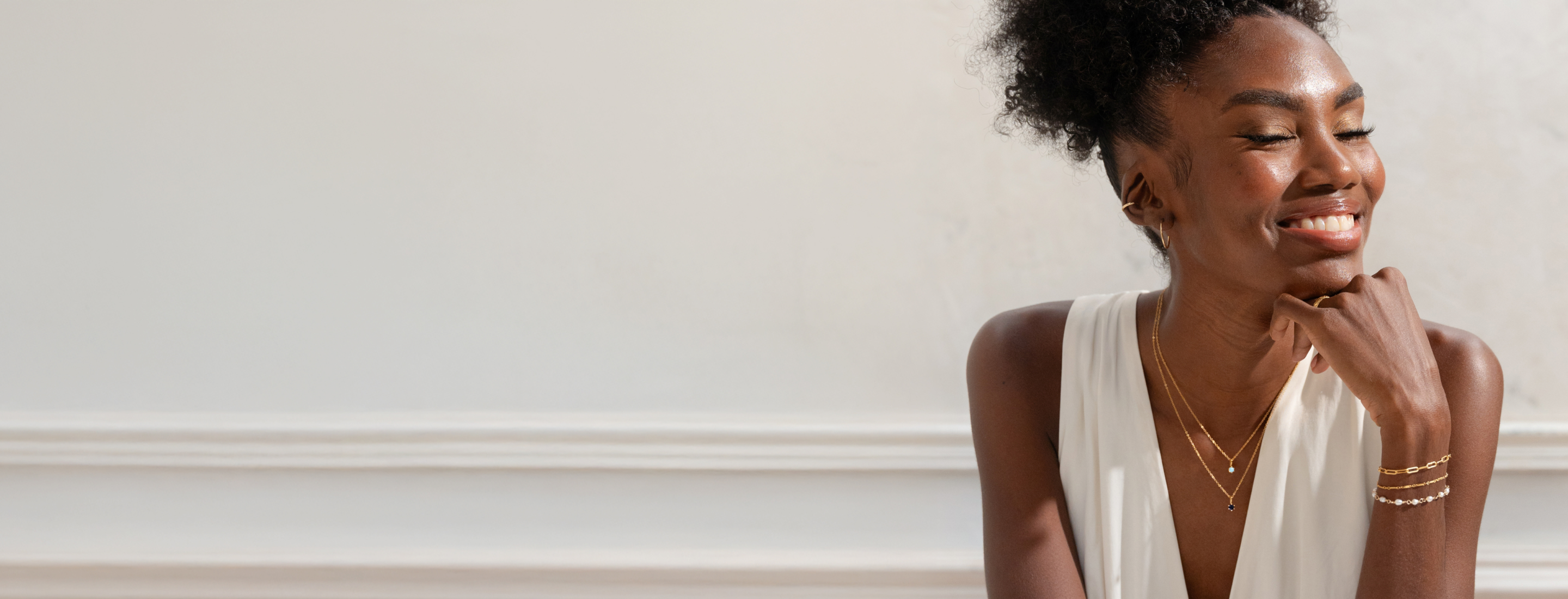Woman smiling with her hand on her chin, wearing layered jewelry including two birthstone necklaces, three bracelets, hoop earrings, and an ear cuff.