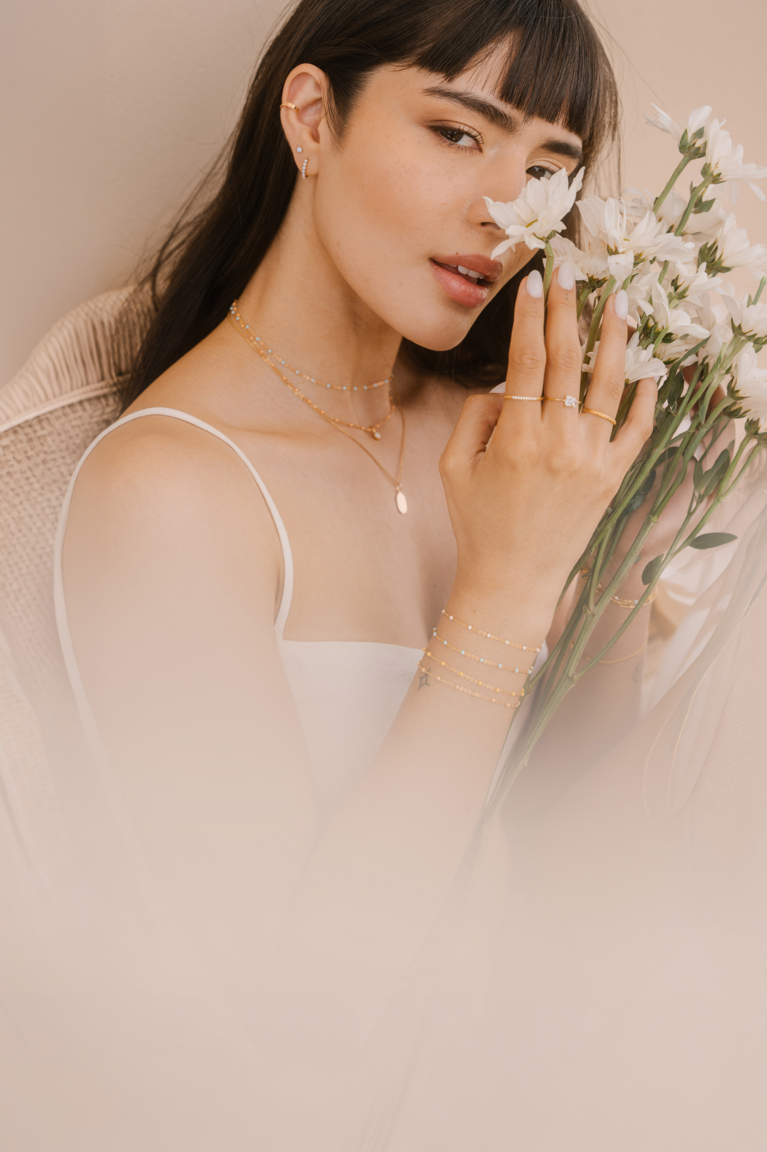 Woman waring gold rings, earrings, and necklaces and holding flowers next to her face.