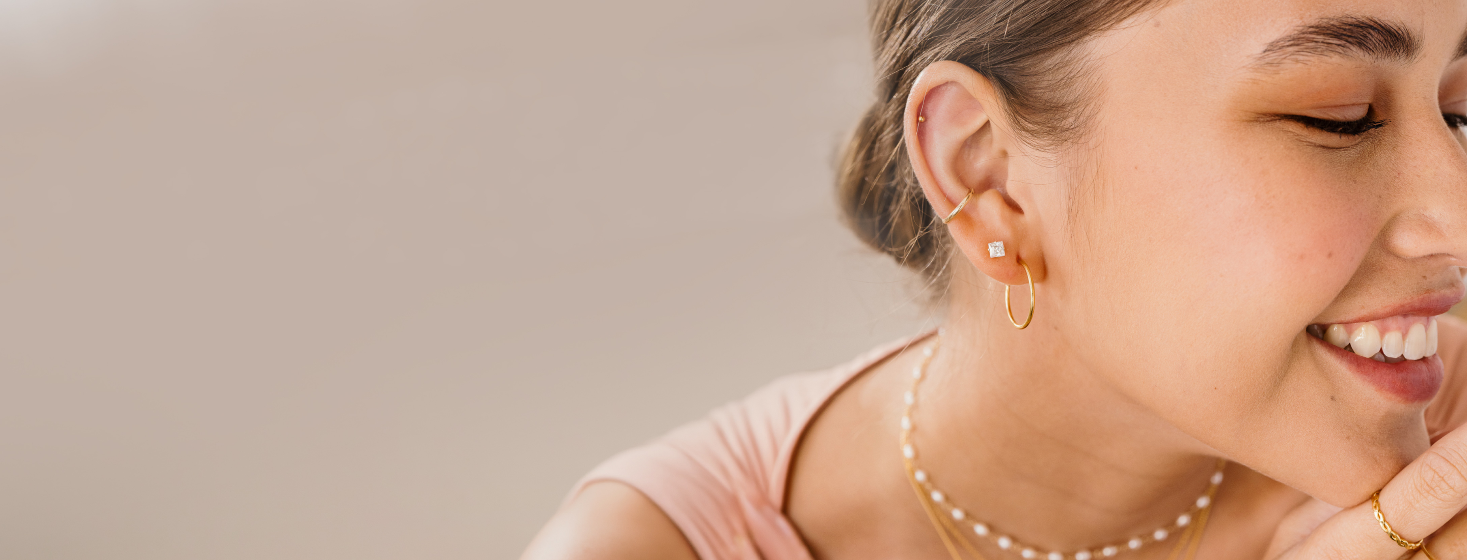 A close-up lifestyle shot of a woman with her hair in a bun, wearing delicate gold hoop earrings, a pearl necklace, and a gold chain against a soft beige background.