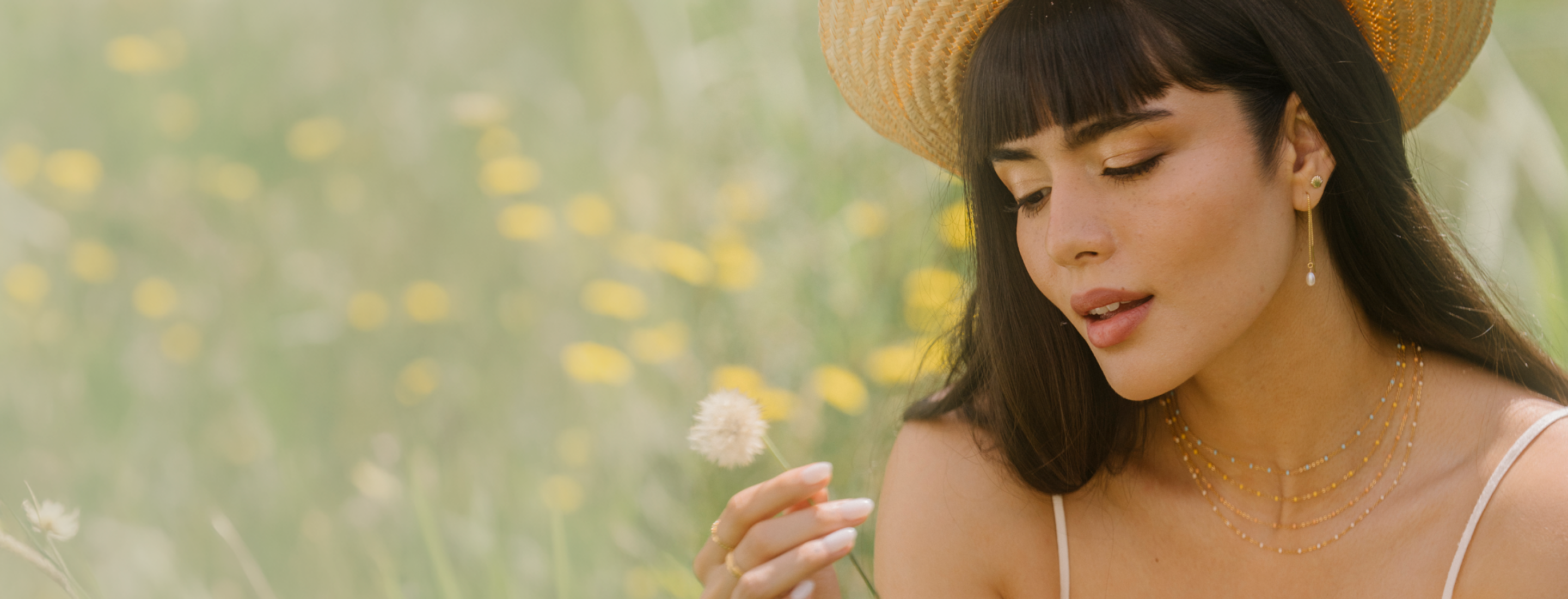 A woman in a straw hat and layered gold enemal necklaces looks down at a dandelion in a sunlit field of yellow flowers.