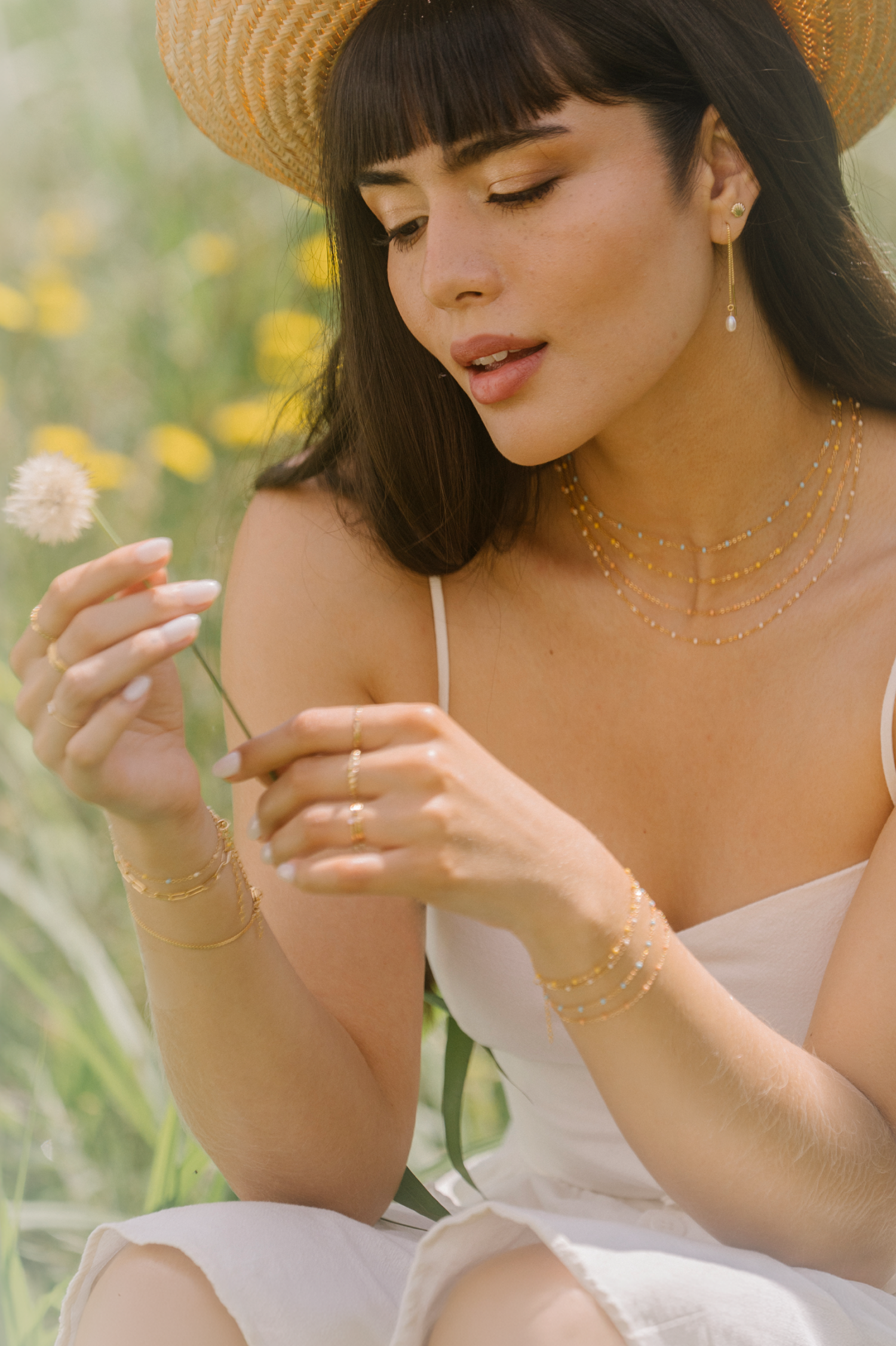 A woman in a straw hat and layered gold enemal necklaces looks down at a dandelion in a sunlit field of yellow flowers.