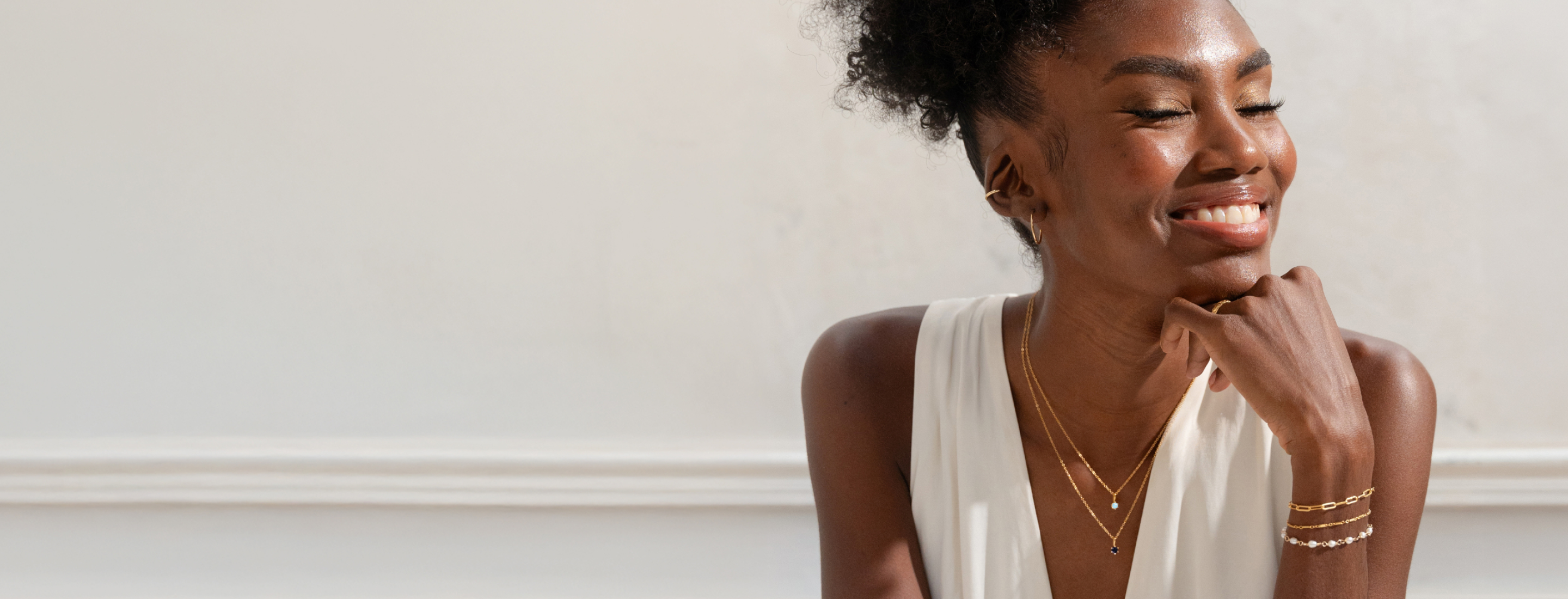 Woman wearing gold jewelry against a white wall. She's wearing 2 birthstone neclaces including a sapphire and an opal birthstone charms. 