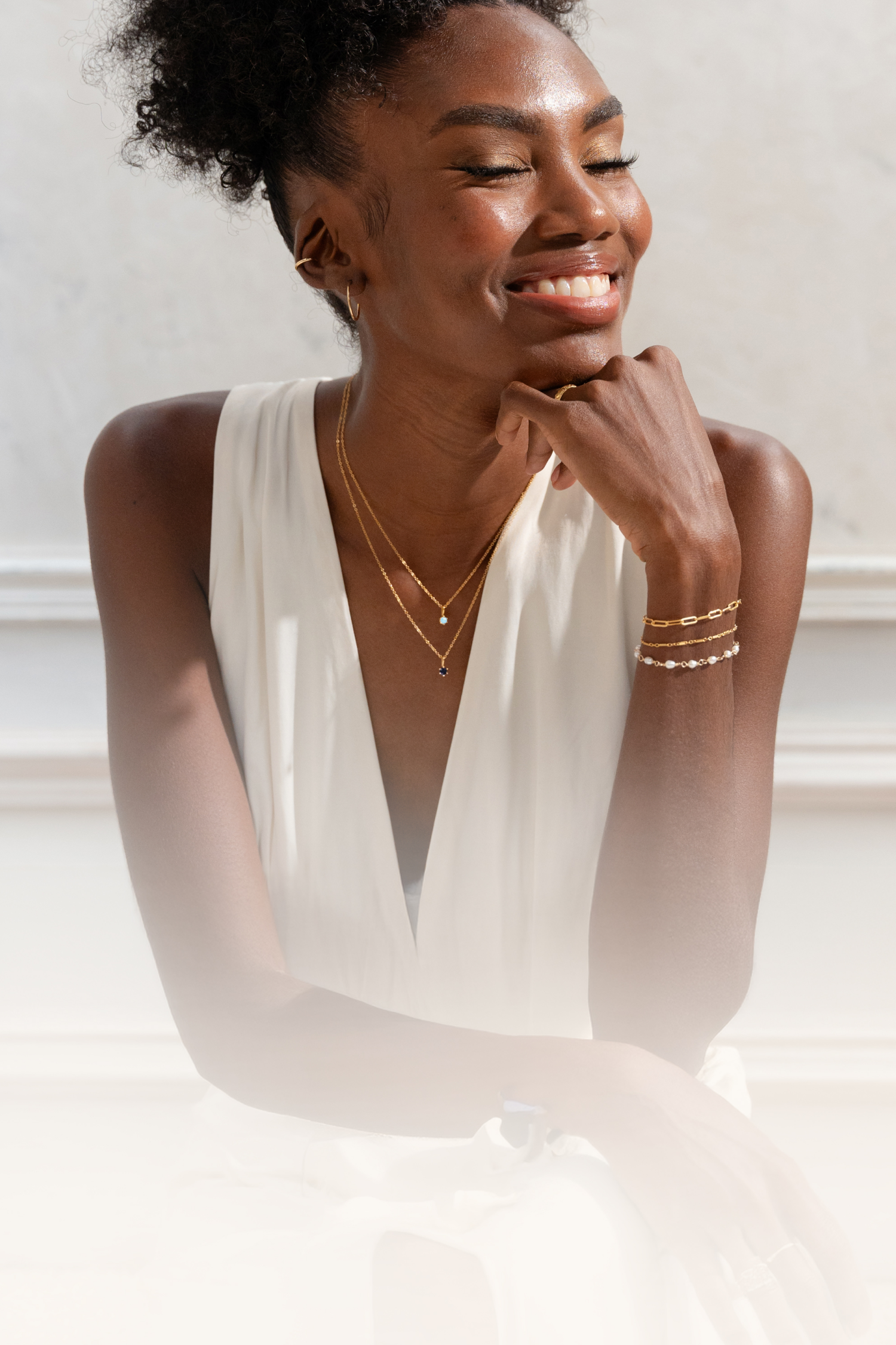 Woman wearing gold jewelry against a white wall. She's wearing 2 birthstone neclaces including a sapphire and an opal birthstone charms. 