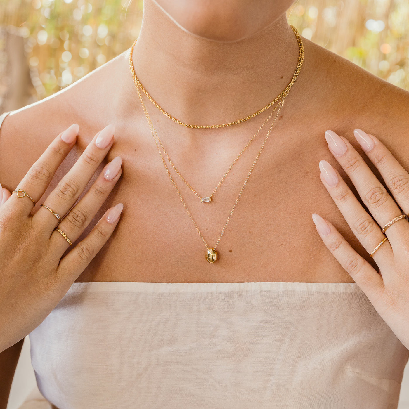 Woman's chest and neck's close up. She's wearing 3 necklaces including rope, dainty baguette, and pebble necklaces and heart knot, dainty baguette, and thin band flower ring on right hand and pavé and rose ring on left hand