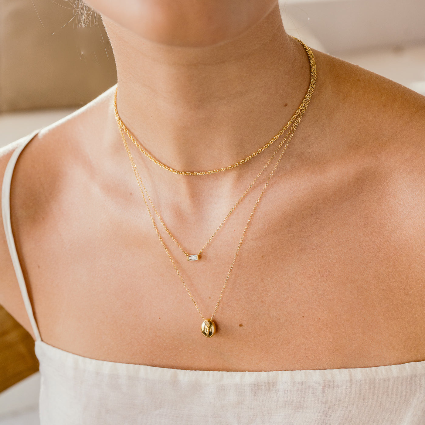 woman's chest and neck close up. She's wearing 3 necklaces including rope, dainty baguette, and pebble necklace.