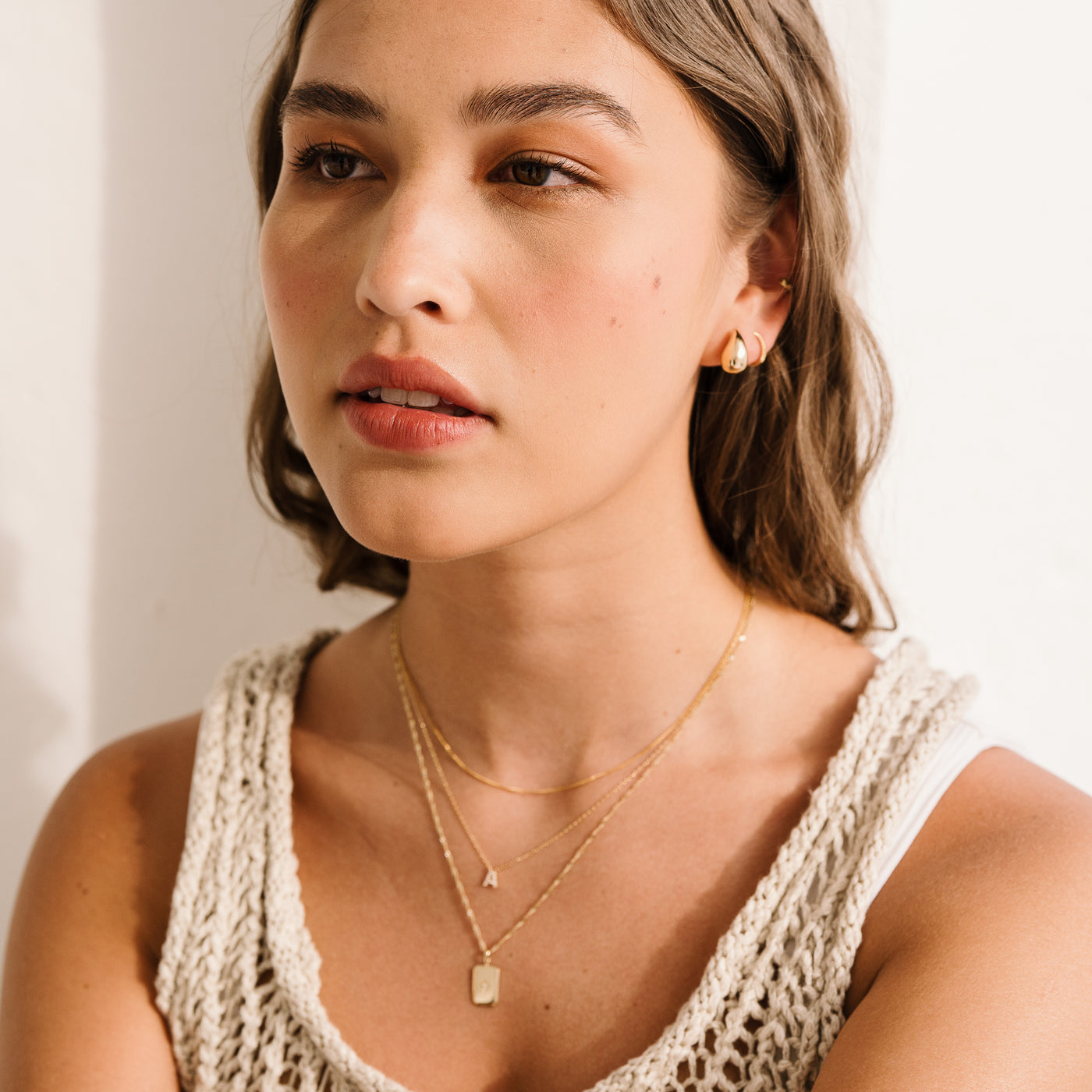 Small Large Teardrop Stud Earrings. Woman in a knitted top wearing 14k gold jewelry including teardrop earrings, hoops, an initial necklace, and a starburst necklace.
