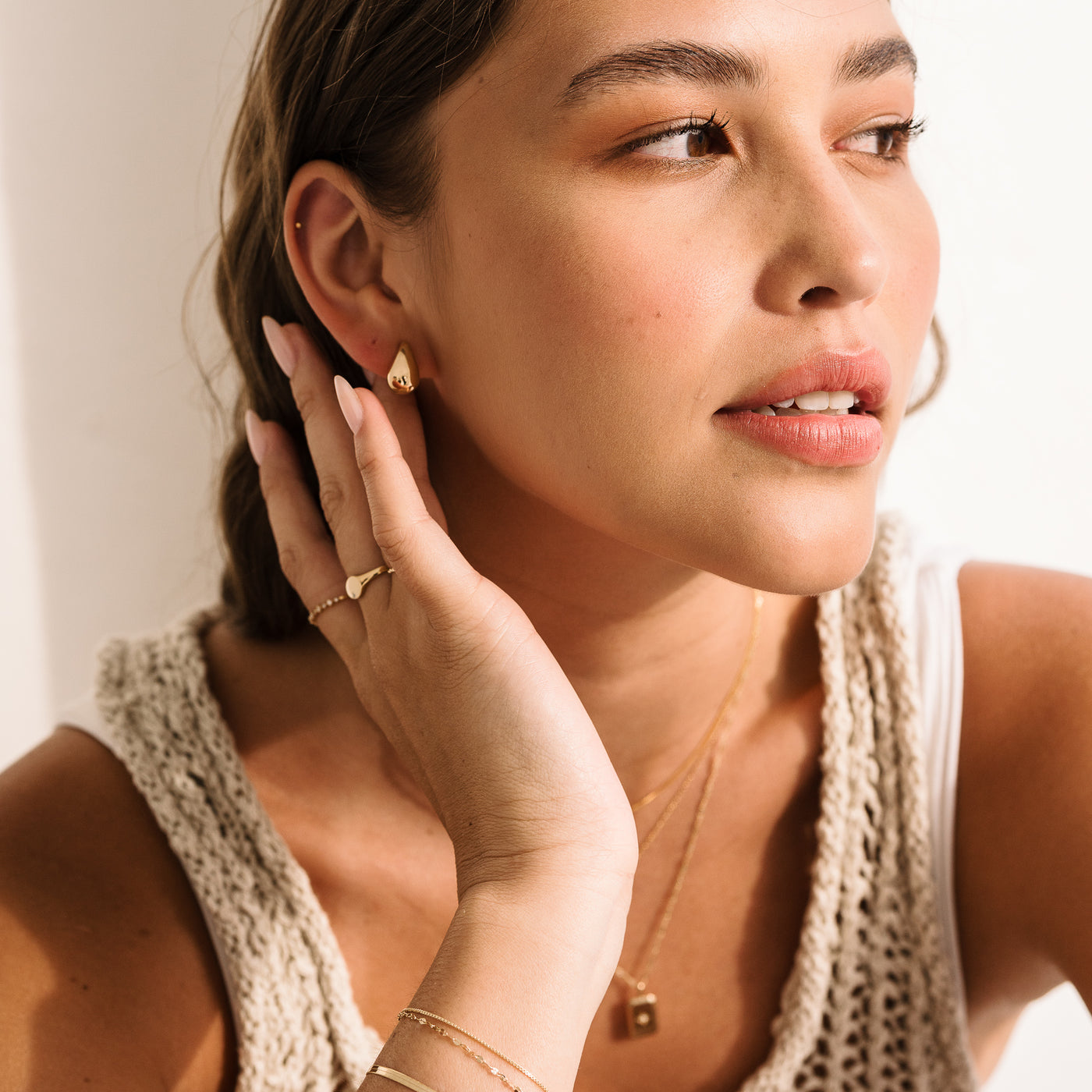 Small Large Teardrop Stud Earrings. Woman looking to the side with her hand near her neck, wearing 14k solid gold jewelry including teardrop earrings, a starburst necklace, a diamond signet ring, and bracelets.