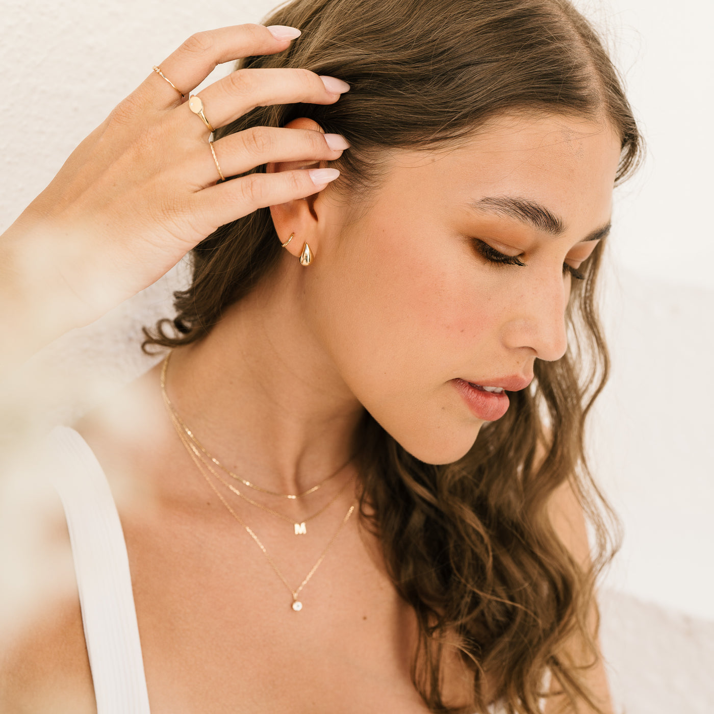 Small Large Teardrop Stud Earrings. woman looking down and tucking her hair under her ear. She's wearing several 14k solid gold jewelry pieces, including gold teardrop earrings, gold hoop, gold chain, initial chain, and diamond bezel necklaces