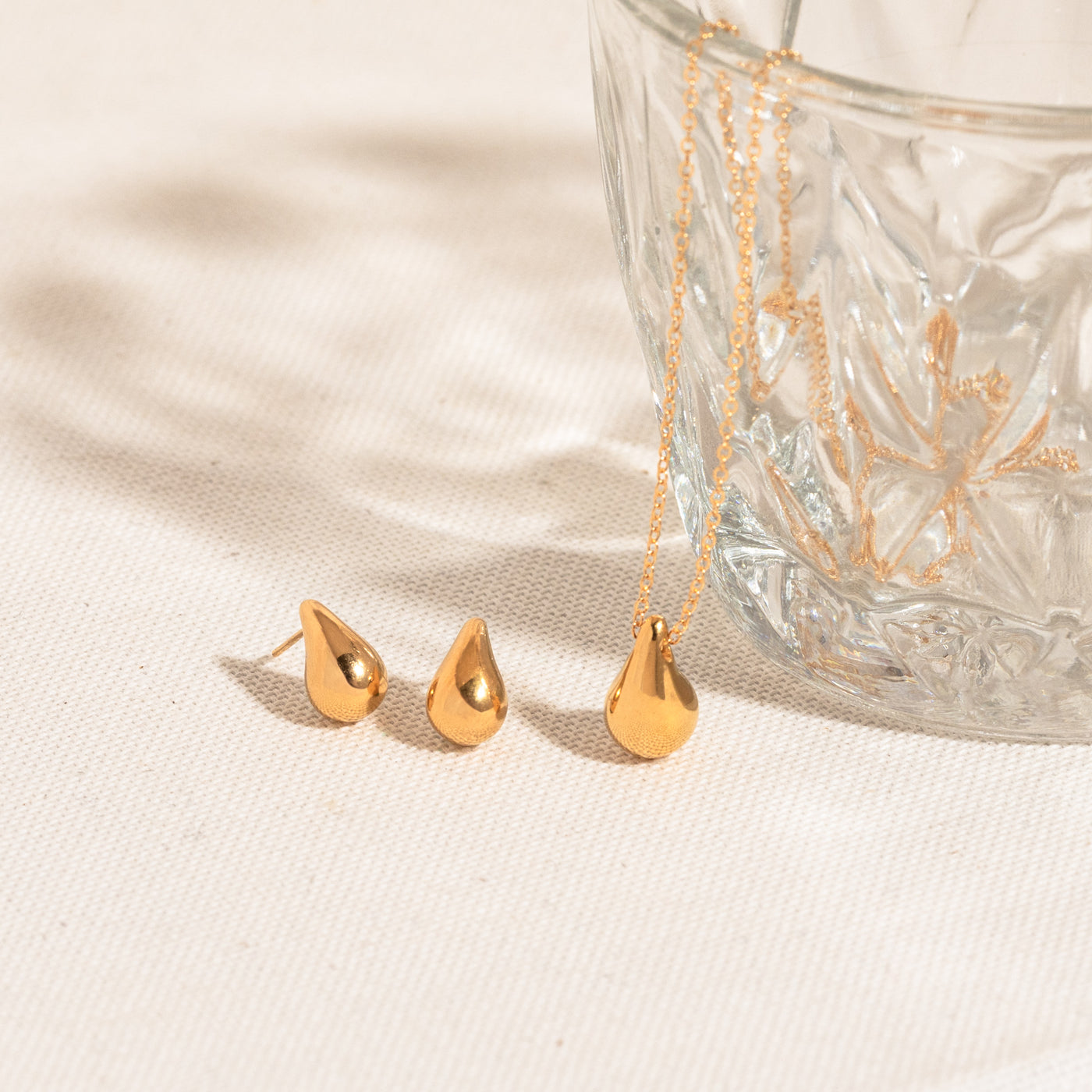 Small Large Teardrop Stud Earrings Dainty teardrop stud earrings and a teardrop necklace lying on a white textured surface, with the necklace chain hanging from a crystal glass.
