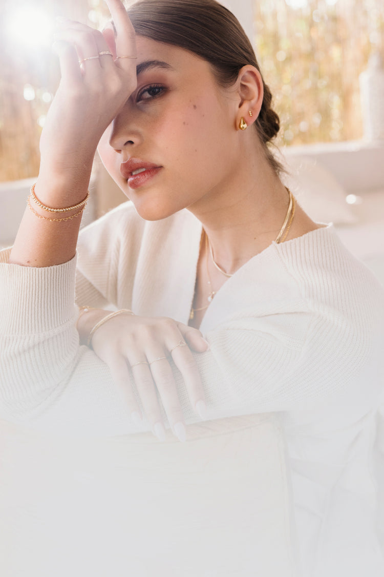 Woman looking to the side wearing teardrop earrings, a gold herringbone necklace, an opal necklace, and a teardrop necklace. She is dressed in a white sweater, with a softly blurred background of natural fiber straws.