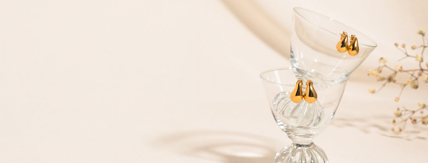 Two pairs of teardrop stud earrings displayed on the rims of stacked glass cups, balanced together. A nude background with small flowers is visible on the right side.