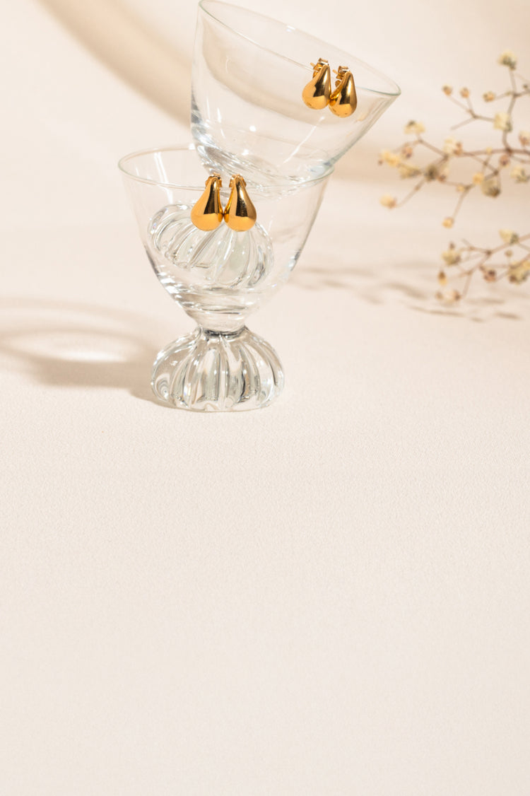 Two pairs of teardrop stud earrings displayed on the rims of stacked glass cups, balanced together. A nude background with small flowers is visible on the right side.