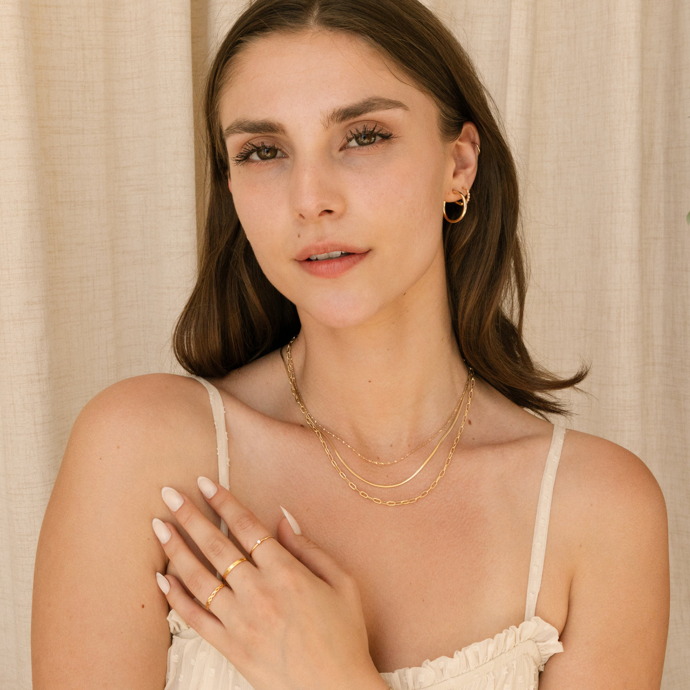Woman in white top wearing 3 chain necklaces, hoop earrings, and rings. Her hand is resting on her chest.