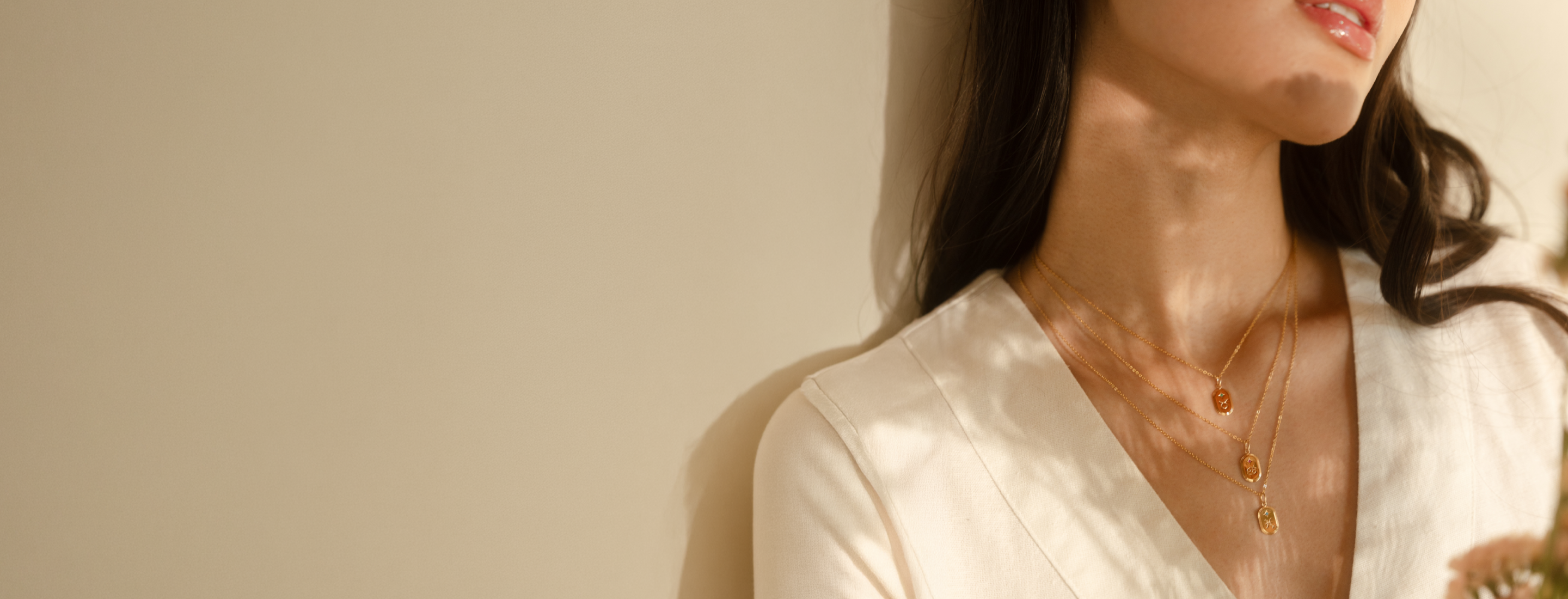 A close-up lifestyle shot of a woman wearing a delicate gold zodiac necklaces. She is wearing a cream-colored top and leaning against a warm beige wall under soft, natural light.
