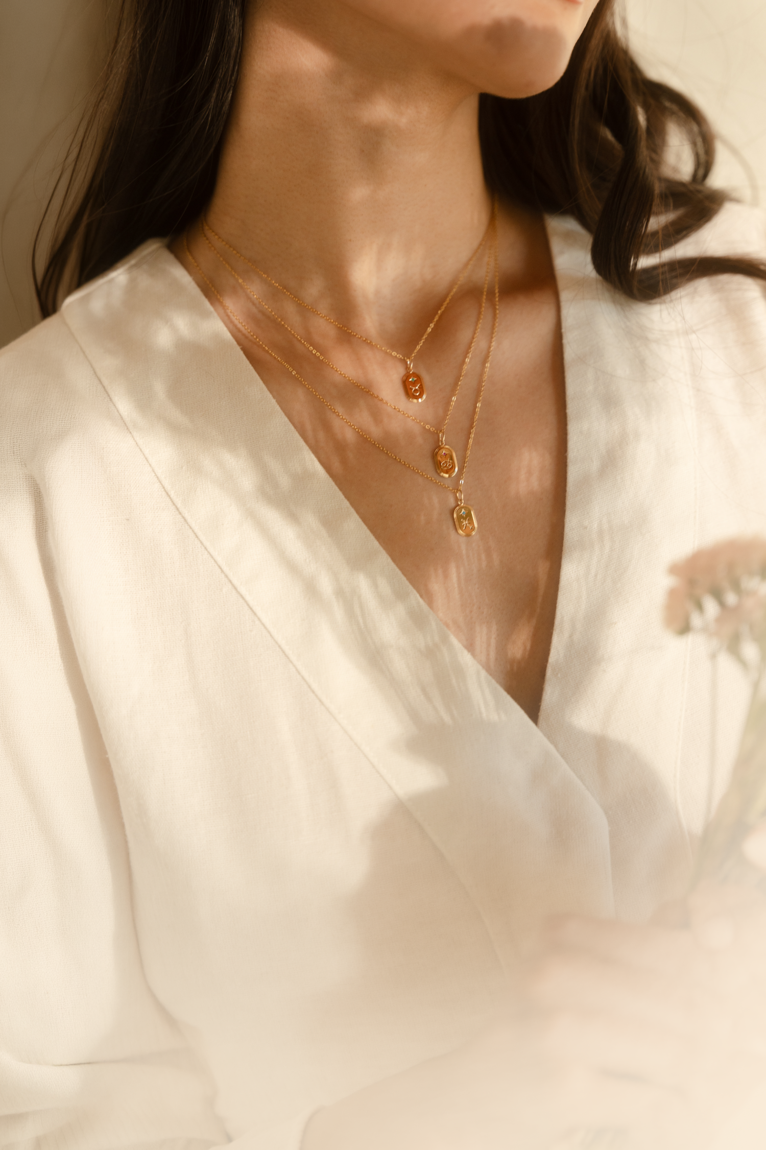 A close-up lifestyle shot of a woman wearing a delicate gold zodiac necklaces. She is wearing a cream-colored top and leaning against a warm beige wall under soft, natural light.