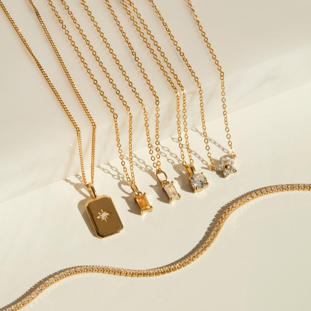 Six gold necklaces: four with CZ stones, one with a citrine stone, and one with a white topaz stone on a beige surface.