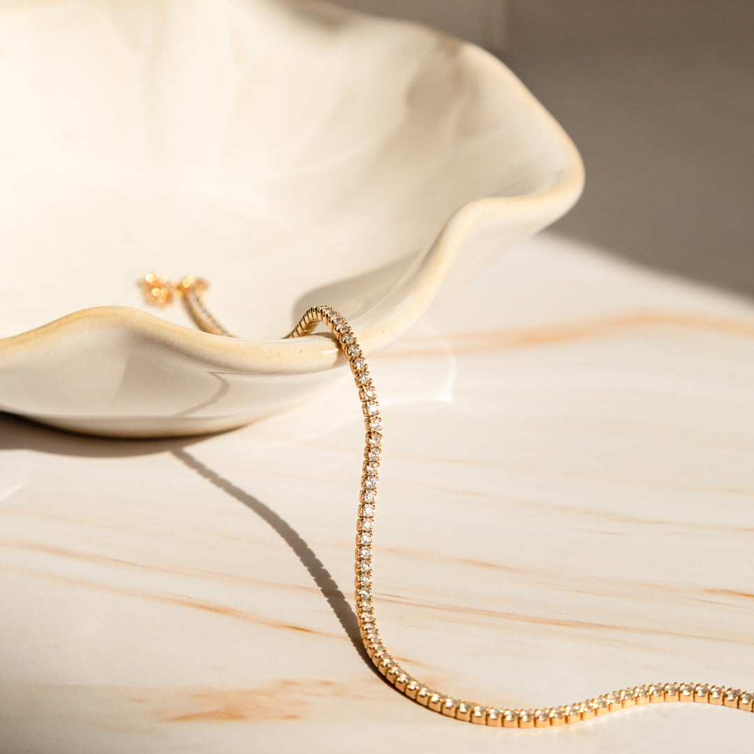 Gold tennis bracelet with cubic zirconia stones draped over a white ceramic bowl on a beige marble surface.